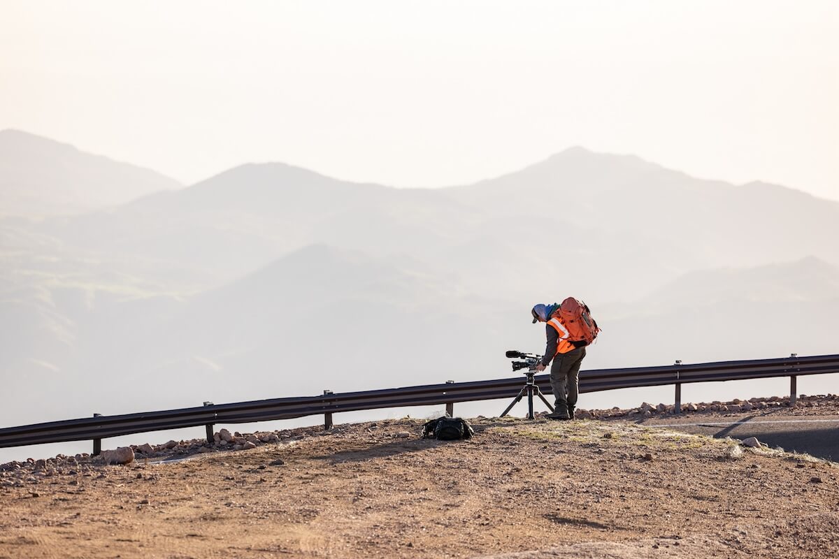 Nick Cahill filming on Pikes Peak. Cameraman with camera, tripod, backpack, and safety vest. Mountain background.